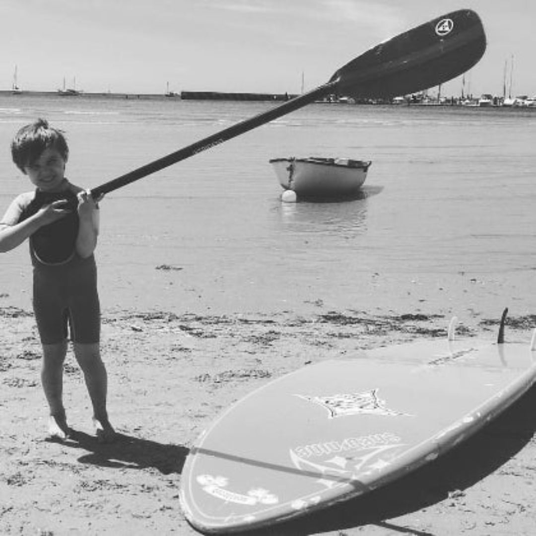 Black and white photo of a child holding a paddle on a beach with a surfboard and boat in the background.