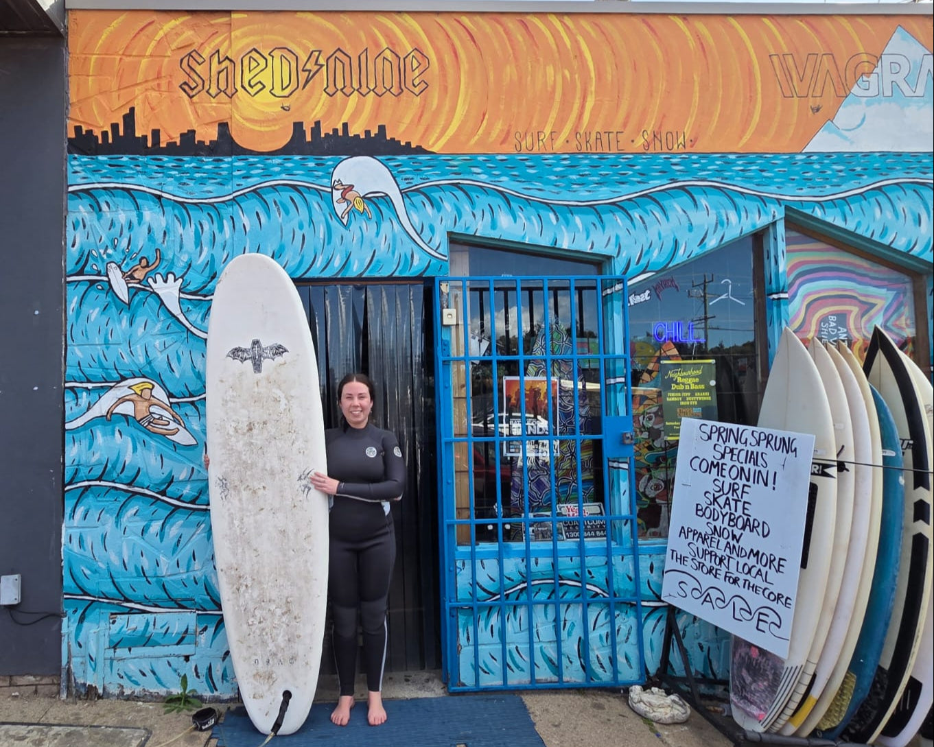 Person holding a surfboard in front of a surf shop named 'Shed Nine' with colorful mural.
