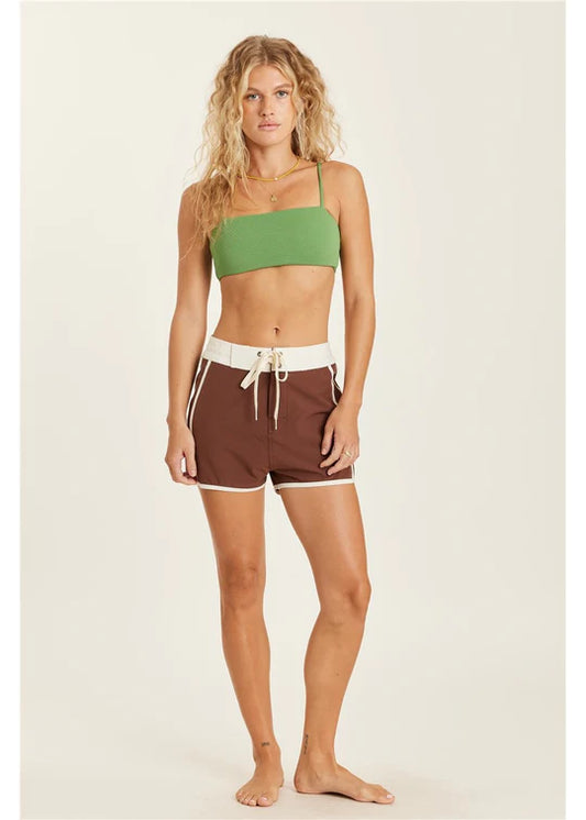 Woman wearing a green bikini top and brown shorts on a beige background shed nine