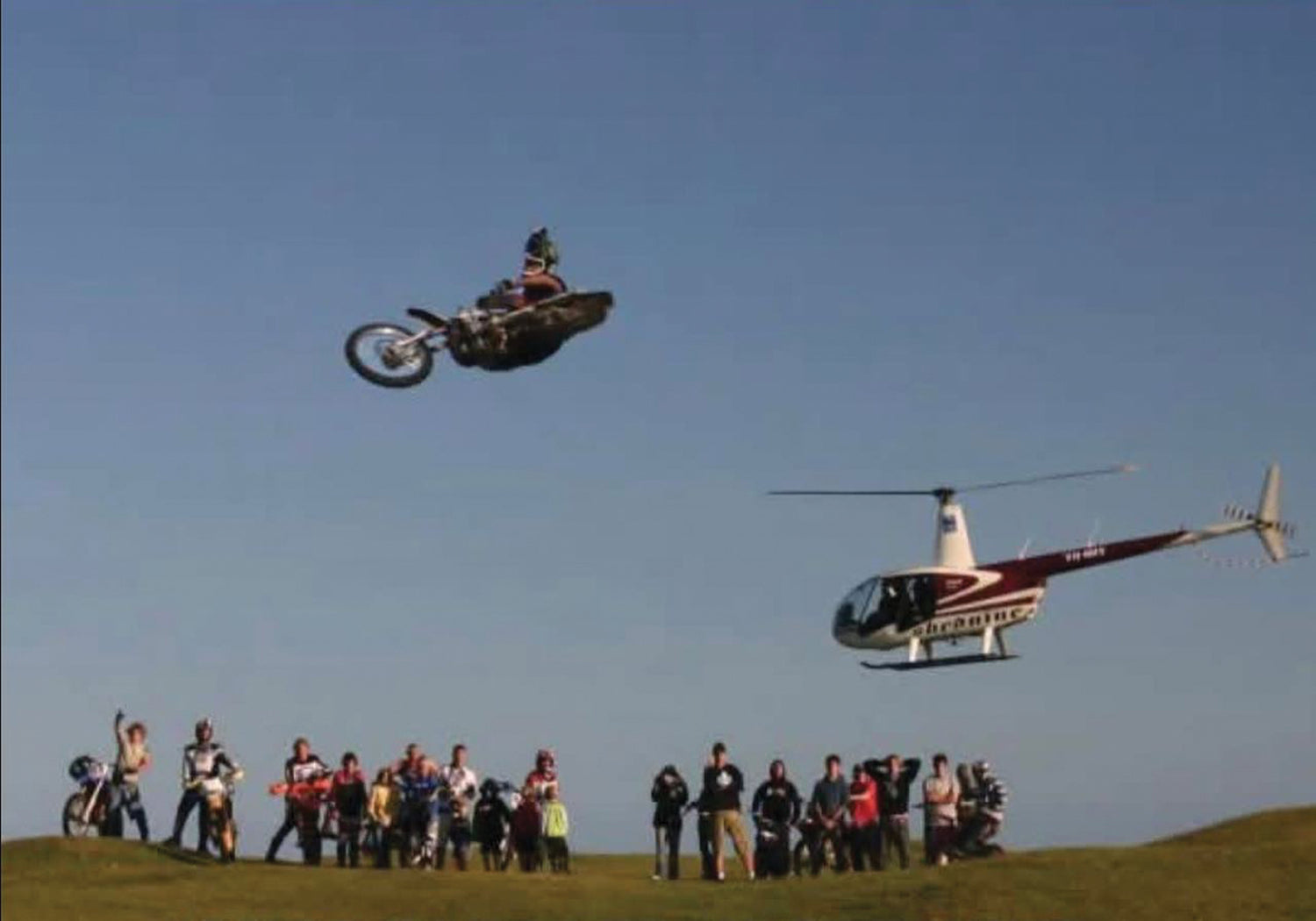 Person on a motorcycle performing a stunt in the air with a helicopter and spectators below.