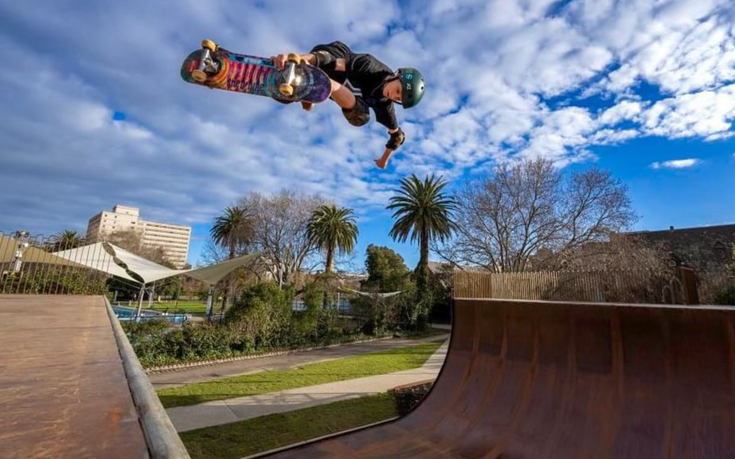 Skateboarder performing a trick over a ramp with a park and buildings in the background