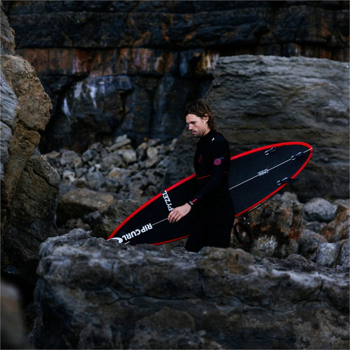 Person in a wetsuit holding a red and black surfboard on a rocky beach shed nine