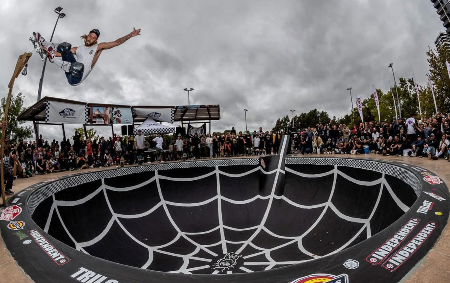 Skateboarder performing a trick in a bowl with a crowd watching on a cloudy day.