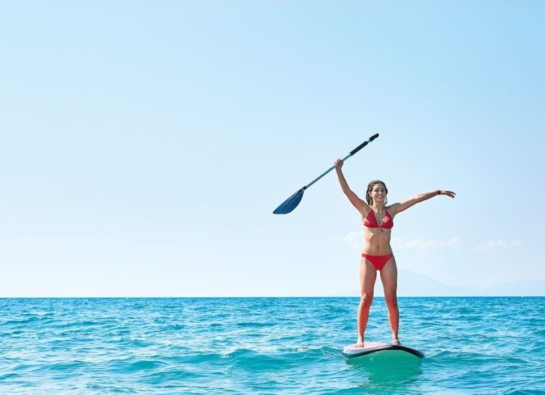 Woman paddleboarding on a clear blue ocean with a blue sky.