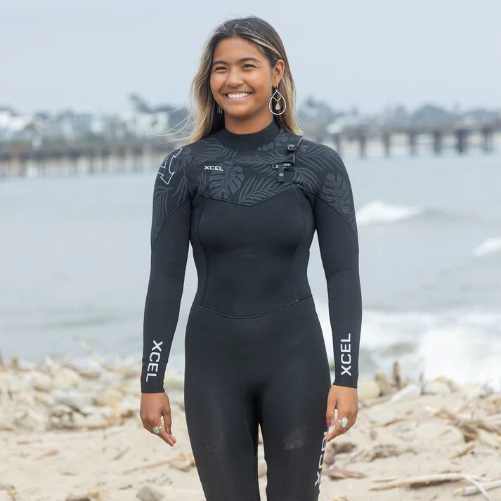 Person wearing a black XCEL wetsuit standing on a beach with water and pier in the background. shed nine