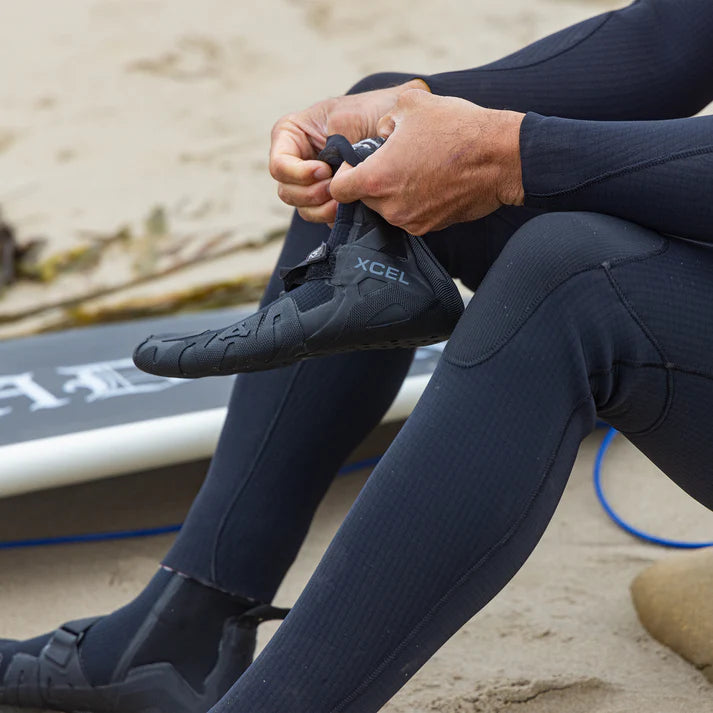 Person in a wetsuit sitting on a beach with a surfboard, tying their XCEL shoe. shed nine