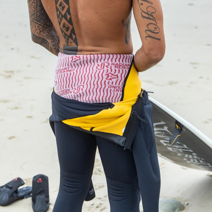 Person wearing a colorful waist bag on a beach with a surfboard in the background shed nine