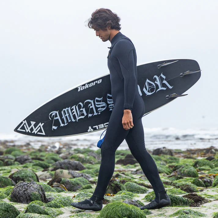Person in a wetsuit holding a Tokoro surfboard on a rocky beach shed nine