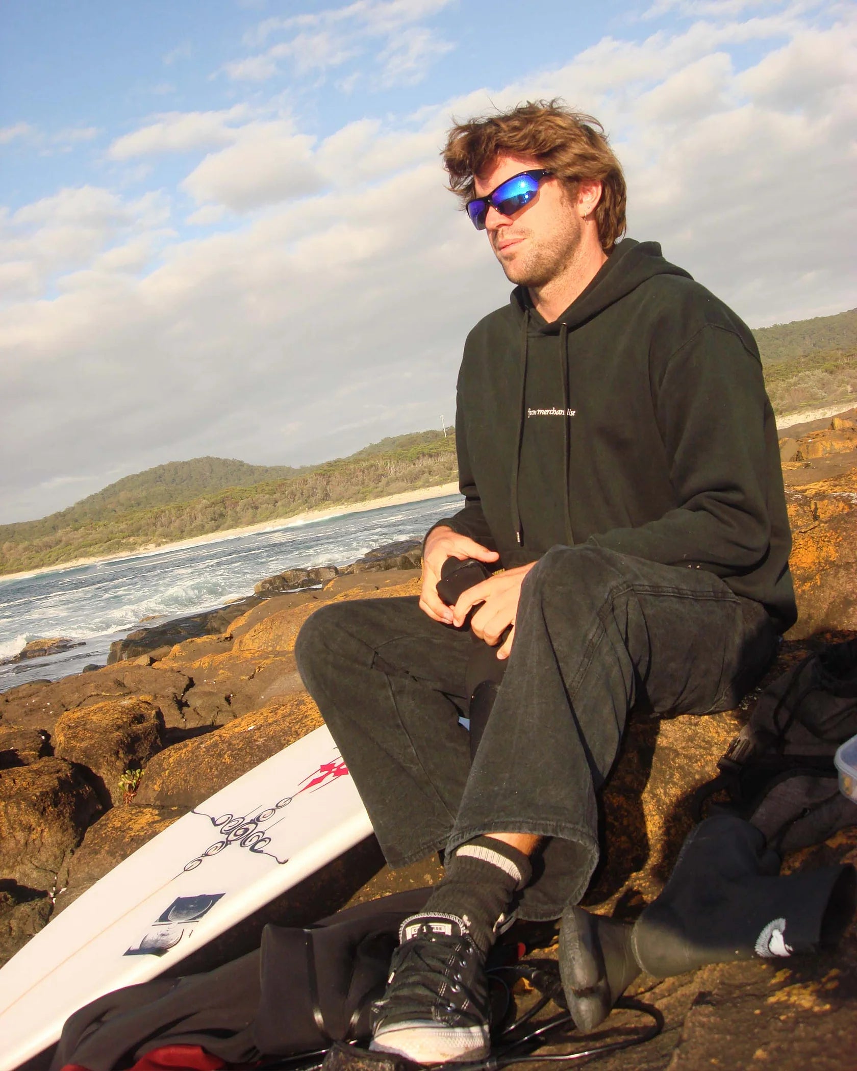 Model wearing Ritual Vision Break & Enter Sunglasses in Polished Black / Reflective Blue seated outdoors with surfboard shed nine