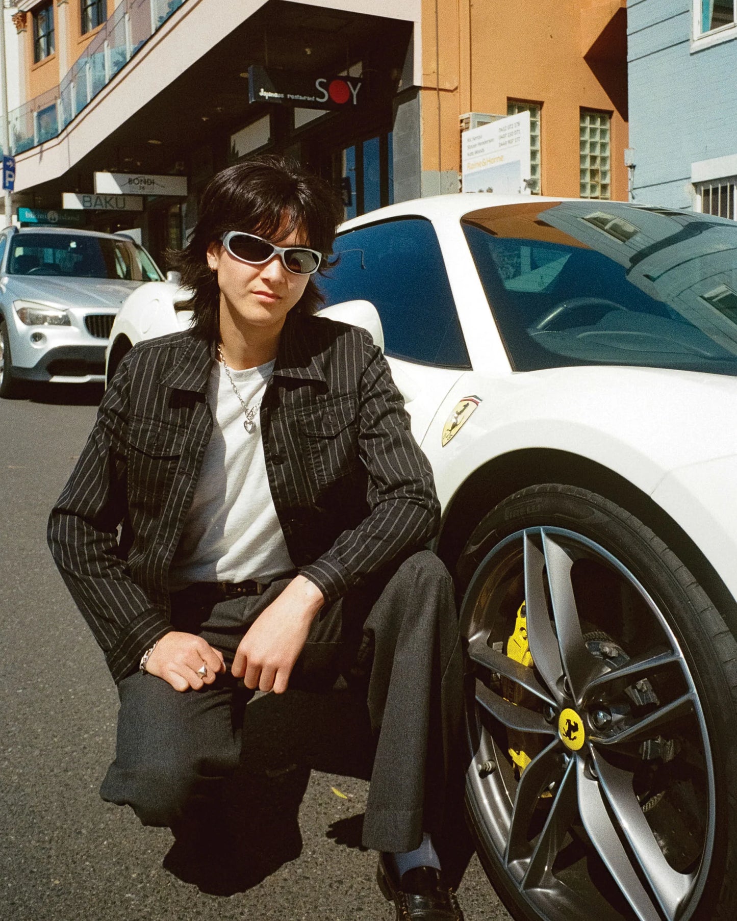 Lifestyle portrait of model wearing Ritual Vision Demon Speed Sunglasses in Matte Chrome / Blue Reflective Polarised 65 mm beside a car shed nine