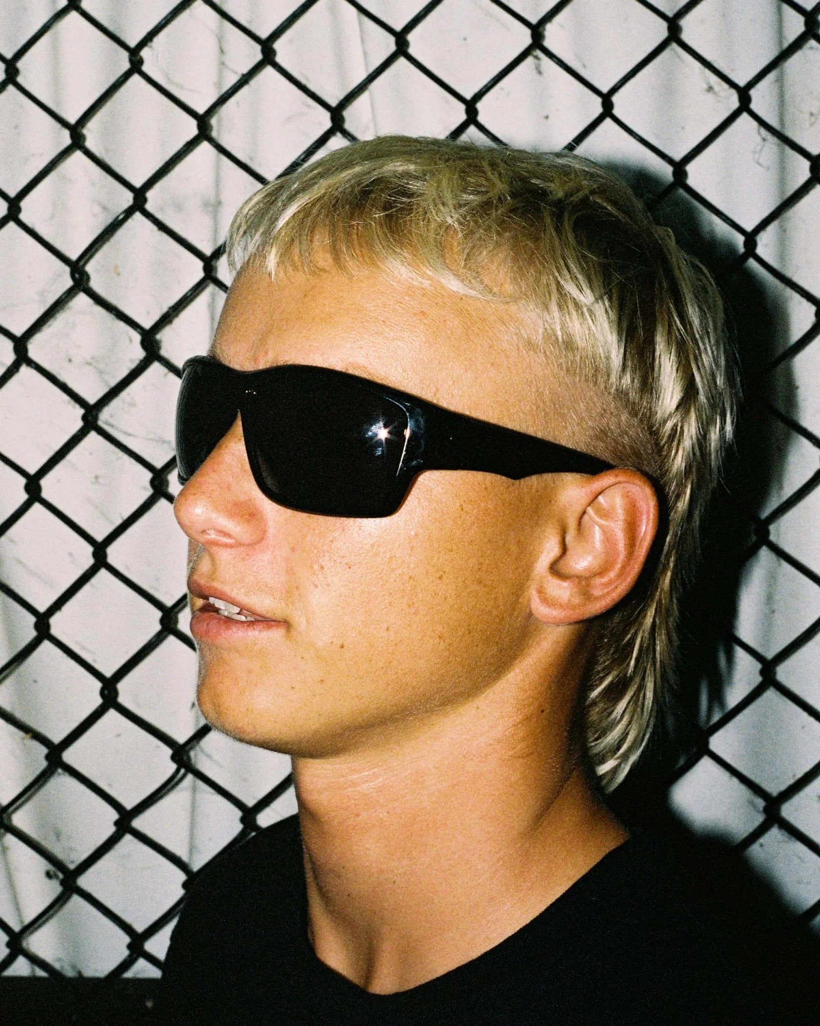 Portrait of model wearing Ritual Vision The Decline Sunglasses in Black / Black Polarised 63 mm by a fence shed nine