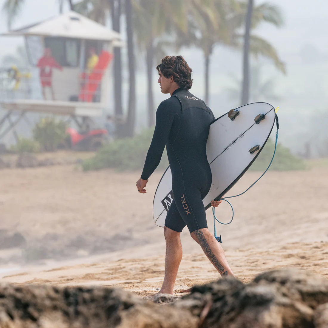 Xcel Men's Comp+ 2mm long-sleeve springsuit in Black, rear-side view on a surfer carrying a board