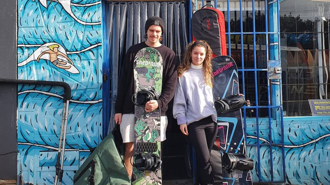 Two people holding snowboards in front of a building with 'Shed Nine' branding.